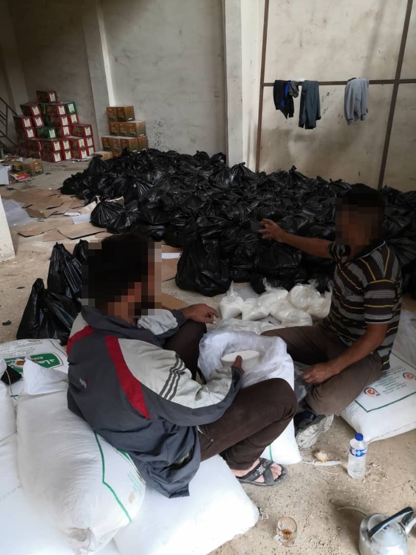 Two people sitting on big packages of rice in a warehouse. In the background are the packages ready for distribution.