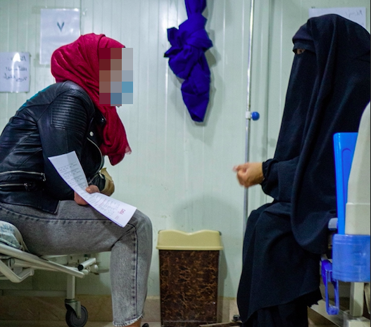 Two women sit across from each other on hospital beds. One woman wears a headscarf, jeans and a leather jacket and holds a patient form in her hand. The other wears a nikab.