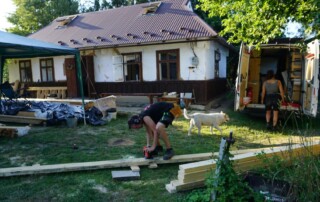 Vor einem alten Wohnhaus werden lange Holzbalken zurecht geschnitten. Neben dem Gebäude parkt ein Transporter, der zu einer Werkstatt umgebaut ist. Vor dem Fahrzeug stehen eine Frau und ein Hund.