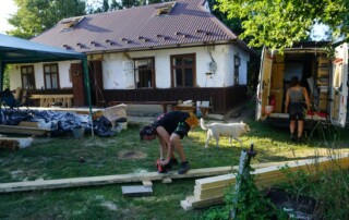 Vor einem alten Wohnhaus werden lange Holzbalken zurecht geschnitten. Neben dem Gebäude parkt ein Transporter, der zu einer Werkstatt umgebaut ist. Vor dem Fahrzeug stehen eine Frau und ein Hund.