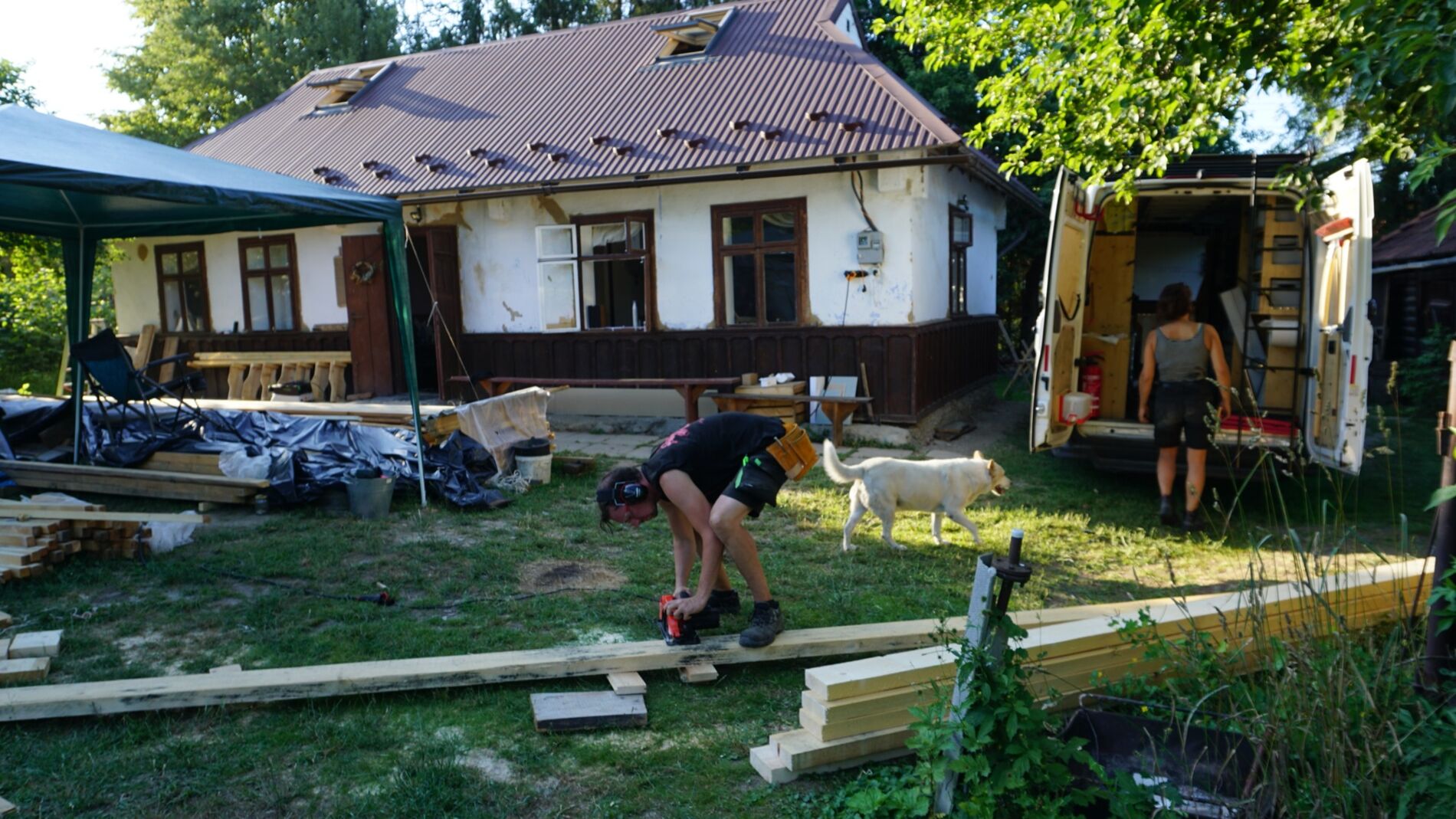 Vor einem alten Wohnhaus werden lange Holzbalken zurecht geschnitten. Neben dem Gebäude parkt ein Transporter, der zu einer Werkstatt umgebaut ist. Vor dem Fahrzeug stehen eine Frau und ein Hund.