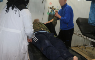 A man lies injured on a stretcher while a second person holds his arm and another person in a white coat stands at his stretcher.