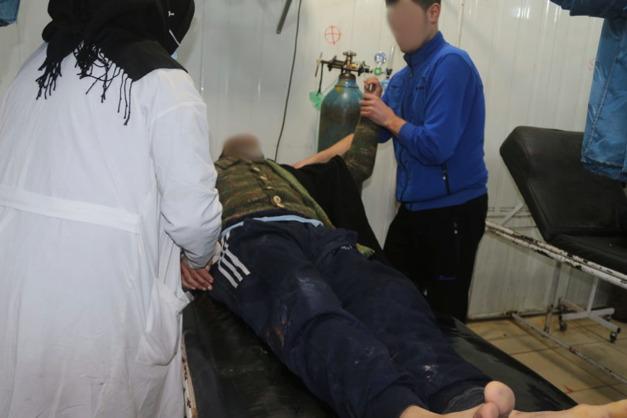 A man lies injured on a stretcher while a second person holds his arm and another person in a white coat stands at his stretcher.