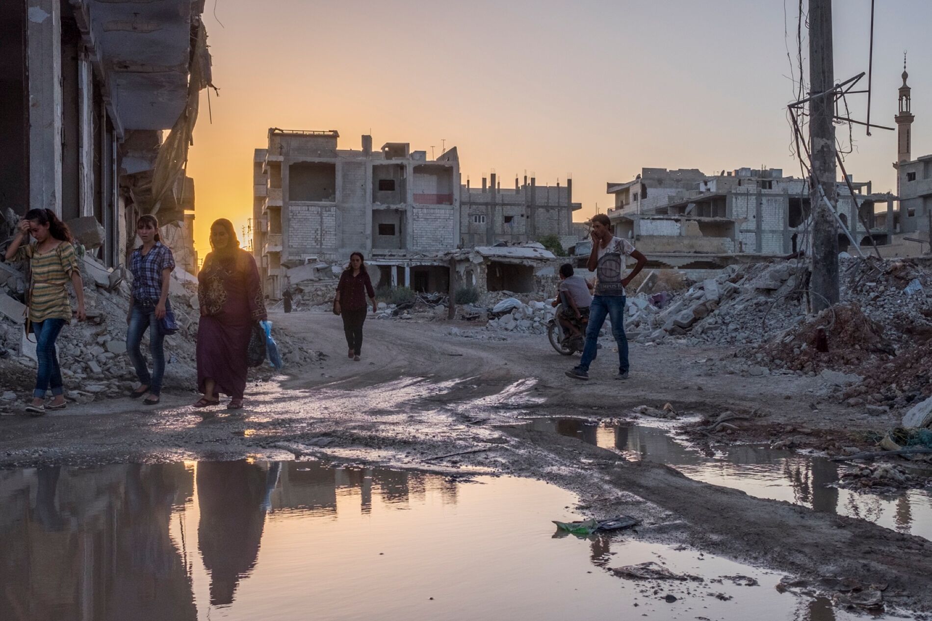 Several people walk across muddy streets in the middle of destroyed buildings.