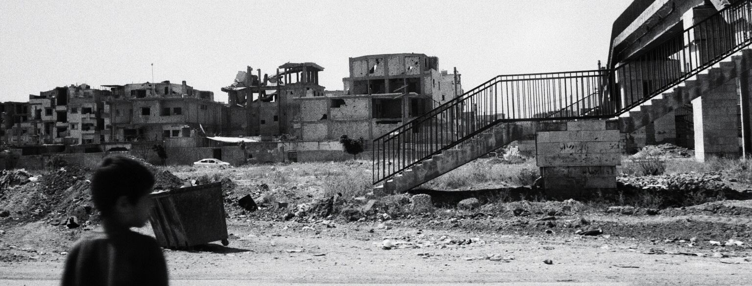 Eine kleiner Junge steht an einer Straße in deren Hintergrund eine Reihe zerstörter Gebäude und eine Brücke sichtbar sind.