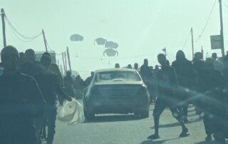Auf einer Straße laufen Menschen links und rechts an einem Auto vorbei in Richtung herabfallender Kisten an Fallschirmen.
